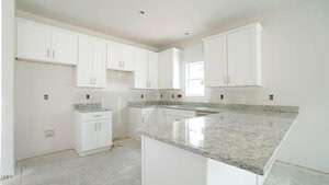 A kitchen under construction features white cabinets, gray granite countertops, and a light gray floor. The walls are painted white, and there is a window above the sink area. Electrical outlets are visible, but the appliances are not yet installed.