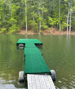 A wooden dock extends into a lake, ending in a green-covered platform. The platform is surrounded by calm water and bordered by a forest of tall, leafy trees.