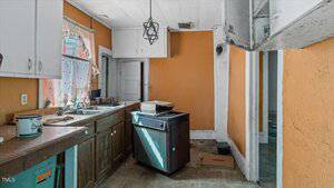 Old kitchen with orange walls, brown cabinets, and a small dishwasher. Counter holds a few items, and a door leads to another room. A pendant light hangs from the ceiling. The room shows signs of wear and some clutter.