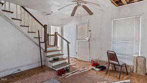 A rundown room with stained carpet and peeling paint. A staircase leads up, and a ceiling fan is missing blades. Blinds cover the windows, and a metal chair sits beside scattered tools. Damaged ceiling panels are visible.