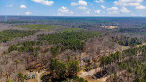 Aerial view of a rural landscape with dense green forests and patches of bare trees. A few houses are scattered along a winding road. The sky is partly cloudy, and vast forested areas extend toward the horizon.