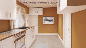 A virtually staged kitchen with white cabinets and wood countertops is shown. The walls are painted orange-brown. A window with beige curtains is on the left and a painting is hung on the right wall. The floor is light-colored wood.