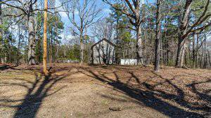 A small, gray wooden house situated in a wooded area with bare trees. The ground is covered in fallen leaves, and there is a clear path leading towards the house. The sky is clear and blue.