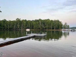 A tranquil lakeside view at 55 Waters Edge Loop in Henderson features a wooden dock extending into the calm water. Two pontoons float serenely on the lake, surrounded by a dense tree line under a partially cloudy sky.
