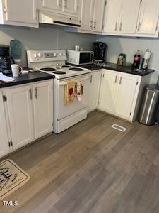 A kitchen with white cabinetry, a black countertop, and a white electric stove. A microwave, coffee maker, and various bottles are on the counter. A rug and trash can are on the wooden floor. Dishtowels hang on the oven handle.