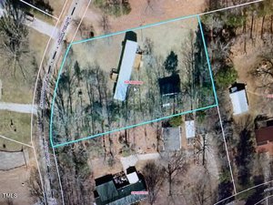 Aerial view of a rectangular property outlined in blue. The area includes a long building, another structure, and several trees. Surrounding roads and neighboring properties are visible.