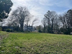 An open grassy field with scattered leaves, bordered by leafless trees under a blue sky stretches across 0 W Young Avenue. A white, two-story house is visible in the background, partially obscured by the trees, capturing the serene essence of Henderson. A small structure sits to the left side of the image.