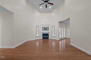 A spacious living room with high vaulted ceilings and a ceiling fan. The room features a fireplace with a mantle, hardwood floors, and two sets of French doors leading to another area. The walls are light-colored.