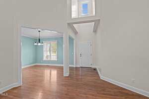 Interior of a house featuring a foyer with a white front door, hardwood floors, and high ceiling. Adjacent room with large window and a white chandelier, painted in a light blue color with white trim. A small window is above the foyer.