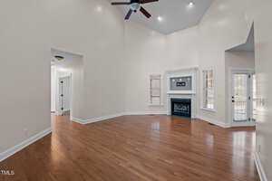 Spacious living room with high ceilings, hardwood floors, and a ceiling fan. The room includes a white fireplace with a mantle, tall windows with blinds, and a door leading outside. Walls are painted in a soft, light color.