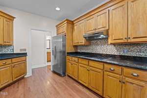 Kitchen with wooden cabinets and hardwood flooring. Features include a stainless steel refrigerator, black countertops, and a backsplash with a mosaic tile design. A stove with an overhead vent is built into the countertop.