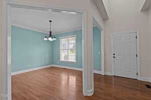 A room with light teal walls and wooden flooring features a window with blinds. There is a three-light chandelier hanging from the ceiling. An open doorway leads to a white door in an adjoining space with similar flooring.