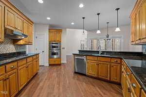 Kitchen interior with wooden cabinets, a dark granite countertop, and stainless steel appliances including a dishwasher, double oven, and range hood. Recessed lighting and pendant lights illuminate the space, which has brown hardwood floors.