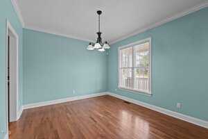 An empty room with light teal walls and a hardwood floor. A window with white blinds is on the right wall, allowing natural light in. A black chandelier with five lights hangs from the white ceiling. A doorway is visible on the left.