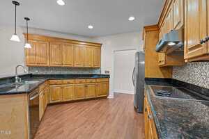 A kitchen featuring wooden cabinetry, stainless steel appliances, and a black granite countertop. A tile backsplash runs along the walls above the counters. The hardwood floor complements the warm tones of the wood finishes.
