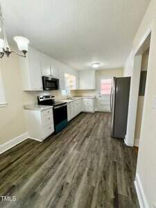 Clean kitchen with white cabinets and black hardware. Features stainless steel appliances, including a stove, microwave, and fridge. Wooden flooring and a chandelier on the left. A window and a back door are visible at the far end.