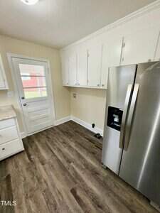 A kitchen with white cabinets, a stainless steel refrigerator, and wood-grain flooring. A small door with a window leads outside. The walls are painted light beige.
