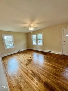 An empty room features polished hardwood floors and cream-colored walls. Two windows let in natural light. A ceiling fan with a light hangs from the center of the ceiling. A closed door is visible on the right side.