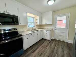 A kitchen with white cabinets, a stainless steel stove, and a black microwave. There's a double sink beneath a window, granite countertops, and dark wood flooring. A door to the right leads outside.