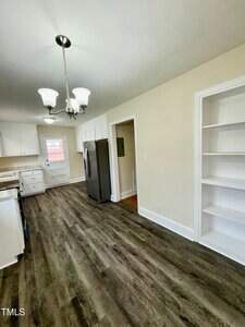 A kitchen with wood-style flooring, white cabinetry, and stainless steel appliances. A built-in bookshelf is on the right wall, and a chandelier hangs from the ceiling.
