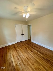 An empty room with wooden floors and beige walls. A ceiling fan with lights is mounted on the ceiling. A closed white double door is on one wall. Natural light enters from an unseen window.