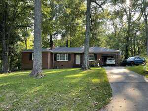 A single-story brick house at 321 Willowood Drive boasts a charming front porch amidst towering trees. Two vehicles rest in the driveway to the right, while a grassy lawn with scattered leaves complements its surroundings. The backdrop features a serene wooded area in Henderson.