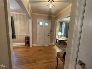 A hallway with wooden flooring and beige walls leads to a front door with a window. A small table is on the right, and a room with a couch and lamp is visible through an archway on the right. A decorative light fixture hangs from the ceiling.