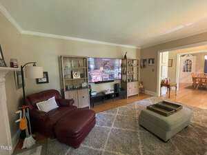 A living room with a large TV centered on a stand between two cabinets. A red leather armchair is next to a white lamp, and a cushioned ottoman sits on a gray rug. The room connects to a dining area with a wooden table and chairs.