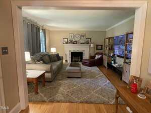 A cozy living room with a gray sectional sofa, a red armchair, and a patterned rug. The fireplace mantel is decorated with a clock and framed photos. A large TV is mounted on a wooden cabinet. The room is well-lit with lamps and natural light.