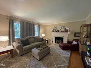 A cozy living room with a large gray sofa and a matching ottoman. A red leather chair and ottoman sit by a white fireplace. The walls feature framed pictures and a large clock. Two lamps and a bookshelf are also present. A soft rug covers the floor.