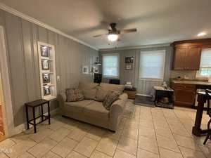 A cozy living room with light gray paneled walls and tile flooring. There's a beige sofa with patterned cushions, a ceiling fan, and a small side table. The room opens to a kitchen area with wooden cabinets and a dog crate on the floor.