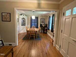 A dining room with a wooden table and six chairs on a rug. There's a cabinet in the corner, large windows with curtains, and walls adorned with framed pictures. A chandelier hangs over the table, and hardwood floors run throughout the room.