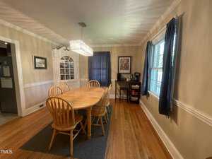A dining room with a wooden table and six chairs on a gray rug. A rectangular chandelier hangs overhead. A white cabinet is against the wall, and a window with blue curtains is on the right. A side table with a coffee maker is nearby.