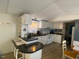 A kitchen with white cabinets, a black countertop, and wood flooring. It features a stove, dishwasher, and a double sink below a window with blinds. A small breakfast bar with two stools is in the foreground, and a dining chair is nearby.
