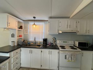 A kitchen with white cabinets, black countertops, and a stainless steel sink. An electric stove, microwave, and coffee maker are on the counters. A small pendant light hangs above the sink, and a window with blinds is behind it. Kitchen items are visible.
