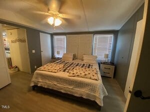 A bedroom with a double bed featuring a plaid and floral duvet, two windows with blinds, and nightstands on either side. A ceiling fan is above, and there's a glimpse of an adjacent room on the left. The walls and floor are neutrally colored.