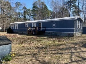 A long, blue mobile home with a small wooden porch, located on a grassy area with dried leaves. Trees and a partly sunny sky are visible in the background. A few yellow flowers are in the foreground.