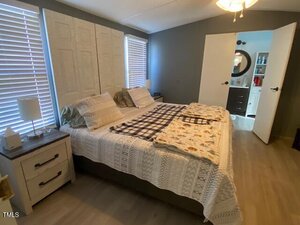 A bedroom with a large bed covered in white and patterned blankets. Two bedside tables with lamps are on each side. The room has gray walls and wood flooring. An open door leads to a bathroom with visible shelves and a mirror.