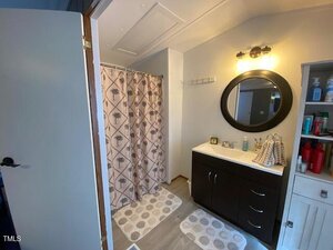 Bathroom with a dark wood vanity and round mirror, illuminated by a wall sconce. A shower with a curtain featuring palm tree designs is on the left. Towels hang on hooks above a cupboard stocked with toiletries. Two patterned rugs cover the floor.