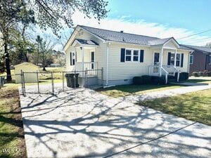 A single-story house with light-colored siding, a few steps leading to a small porch, and a gabled roof. There is a concrete driveway, and a chain-link fence with a gate. Trees and a shed are visible in the background.