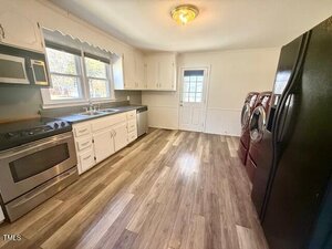 A kitchen with wood flooring, featuring a stove, microwave, and sink on the left. A washer and dryer stand on the right near a large black refrigerator. Cabinets are above and below the countertops. A door at the back leads outside.