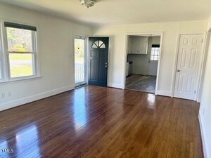 A bright, empty living room with polished hardwood floors and white walls. There are two windows on the left, a dark blue front door, and openings leading to a kitchen and another room. Sunlight illuminates the space through the windows.