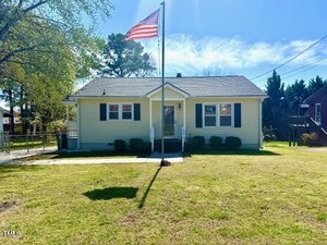 A yellow single-story house with a gray roof sits serenely at 509 Sunset Avenue in Oxford. An American flag flies proudly in the front yard, where trimmed bushes line the house and a large tree stands to the left under a clear blue sky.