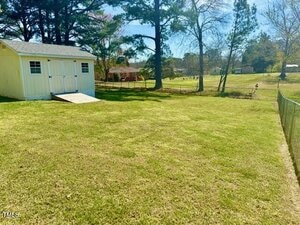A well-maintained backyard with a green lawn and a light yellow shed with white double doors. The yard is bordered by a metal fence on one side. In the background, there are trees and some outbuildings. The sky is clear and sunny.
