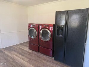 A laundry room with two front-loading red washing machines and a large black refrigerator with a water and ice dispenser. The floor is wood-textured, and the walls are light-colored.