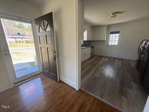 View of an entryway with a wooden floor leading to a kitchen area. The kitchen has light-colored cabinetry, a dark countertop, and a door leading outside. A washing machine and dryer are visible to the right. Natural light illuminates the space.
