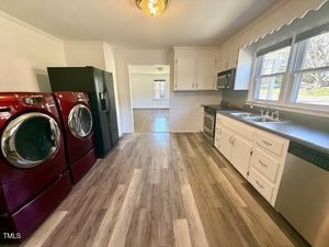 A laundry room with red front-loading washer and dryer on the left, white cabinets along the right wall, a black countertop, and a sink under a double window. The floor is wood-patterned, and there's a glimpse of an adjacent room in the background.