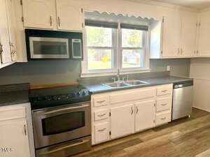 A kitchen featuring white cabinets and gray countertops. There is a stainless steel oven with a gas stovetop, a microwave above it, and a dishwasher to the right. A double sink with a window above is centered on the back wall.