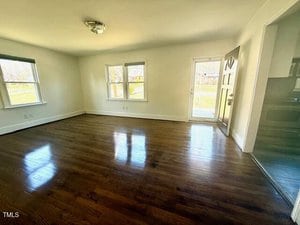 An empty room with shiny, dark hardwood floors and white walls. Three windows on the far wall provide natural light. An open door leads outside, and a kitchen area is partially visible on the right. A ceiling light fixture is centered above.