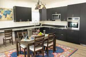 A modern kitchen with dark cabinets, stainless steel appliances, and a mosaic light fixture. A wooden dining table with four chairs is placed on a colorful rug. A wall map and window add decorative touches.